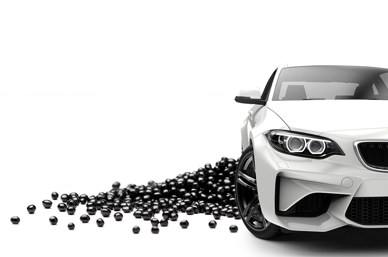 Black shiny polymer pellets are laid on white background, in front of it, a white sports car is placed 